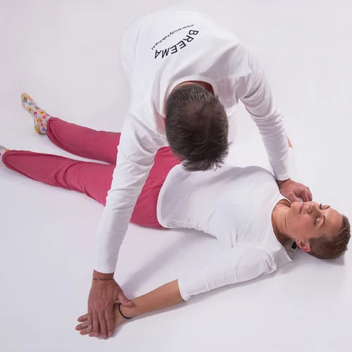 Two practitioners performing a close floor-based bodywork exercise with focused attention.