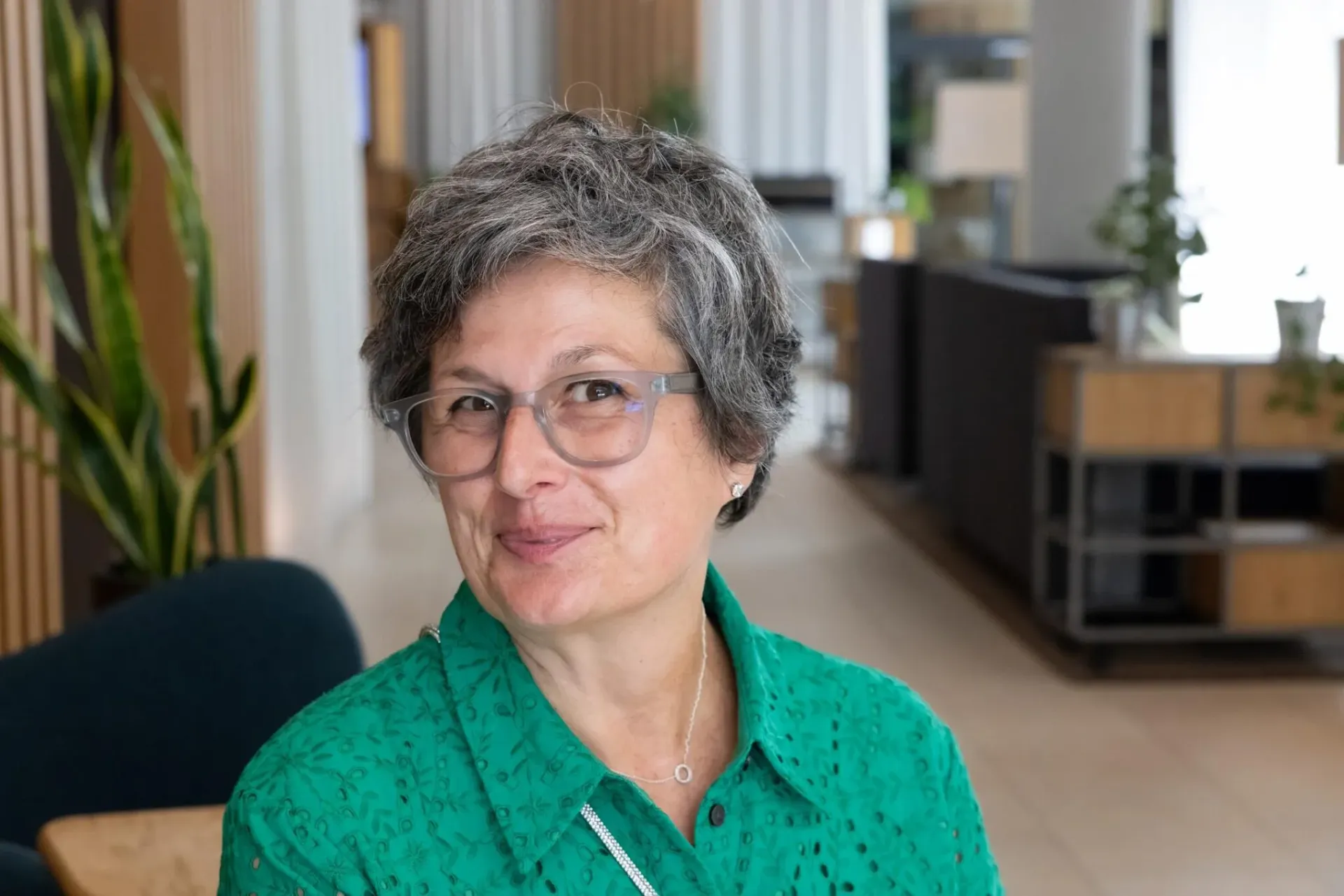 Woman with short grey hair and green eyelet blouse smiles warmly in a modern office setting.