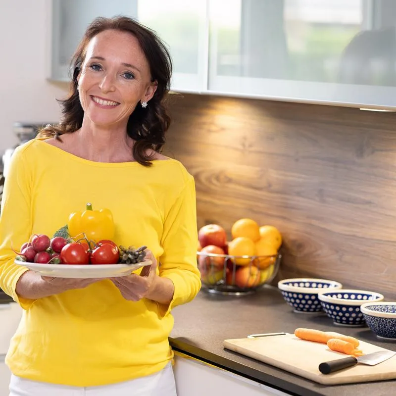 Woman in yellow top holds a plate of colorful vegetables and peppers in kitchen.