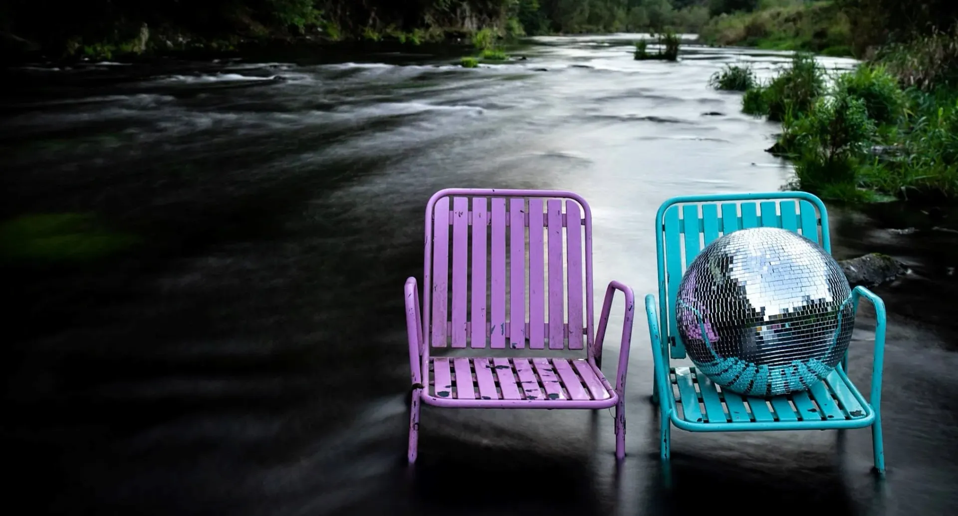 Two vintage chairs beside a flowing river, one holding a disco ball, evoking reflective tranquility.