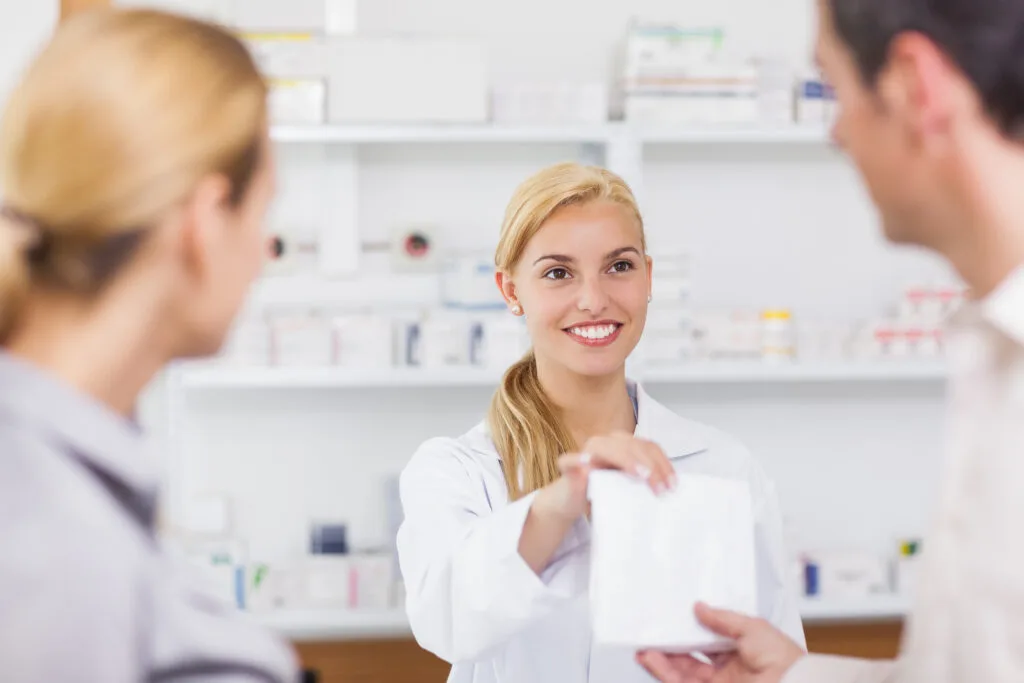 A smiling pharmacist in a white coat handing medication to customers, perfectly embodying the warm, personal consultation at Diana Apotheke.