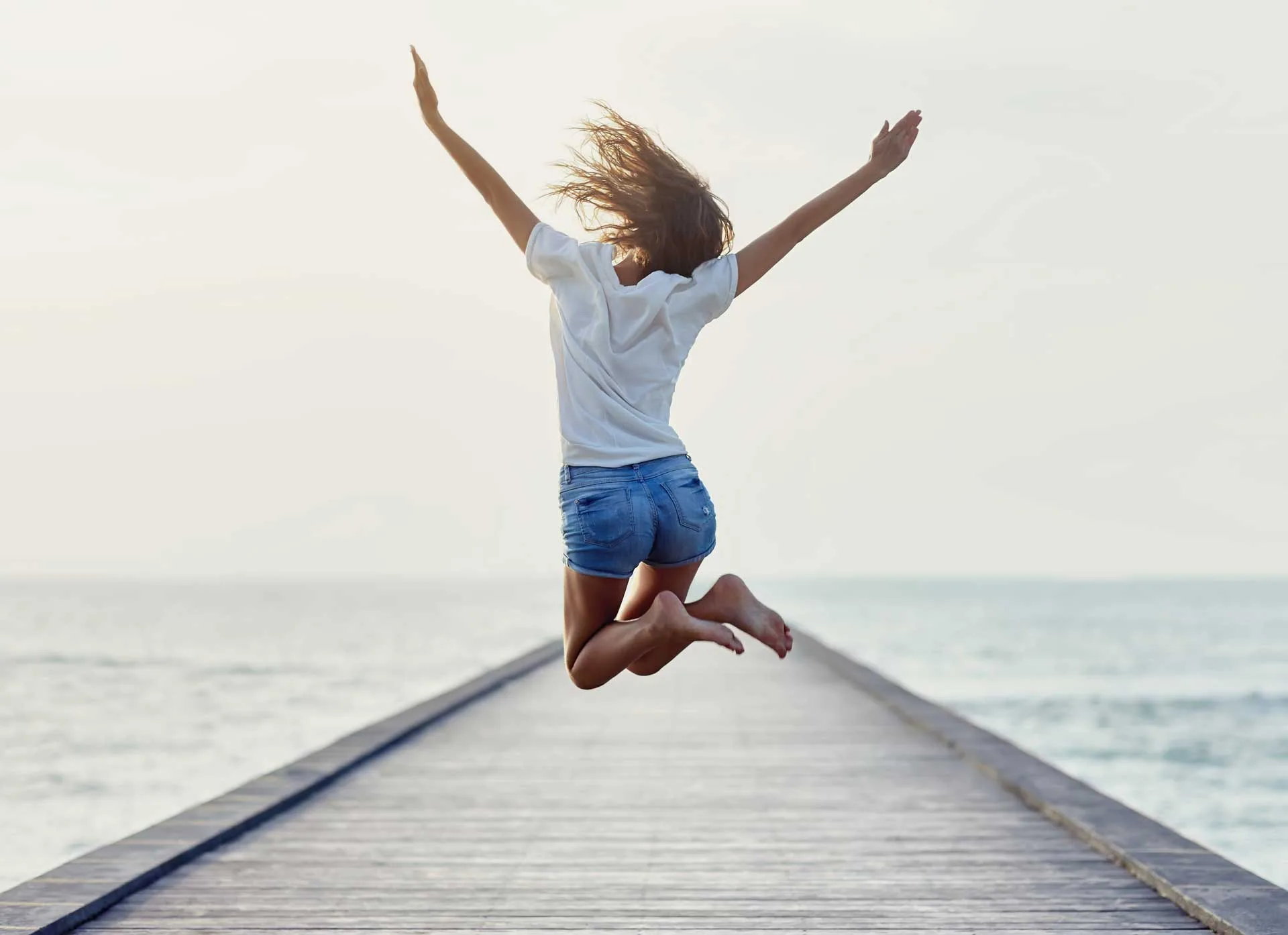 A joyful woman leaps with arms raised on a sunlit ocean boardwalk.